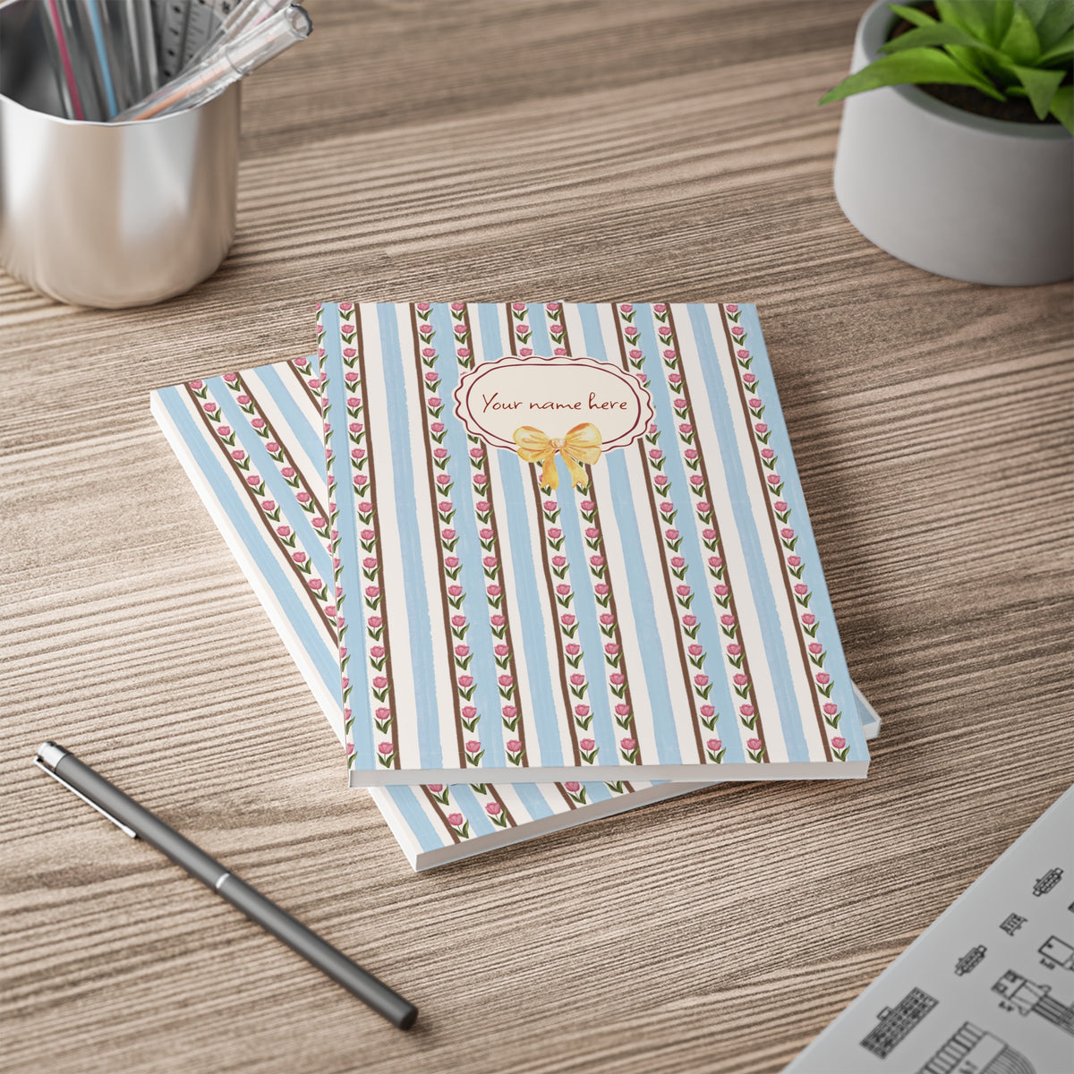 Two personalized sofctover notebooks A5 with a light blue and brown floral striped cover, stacked on a wooden desk next to a pen, with a plant and cup in the background.