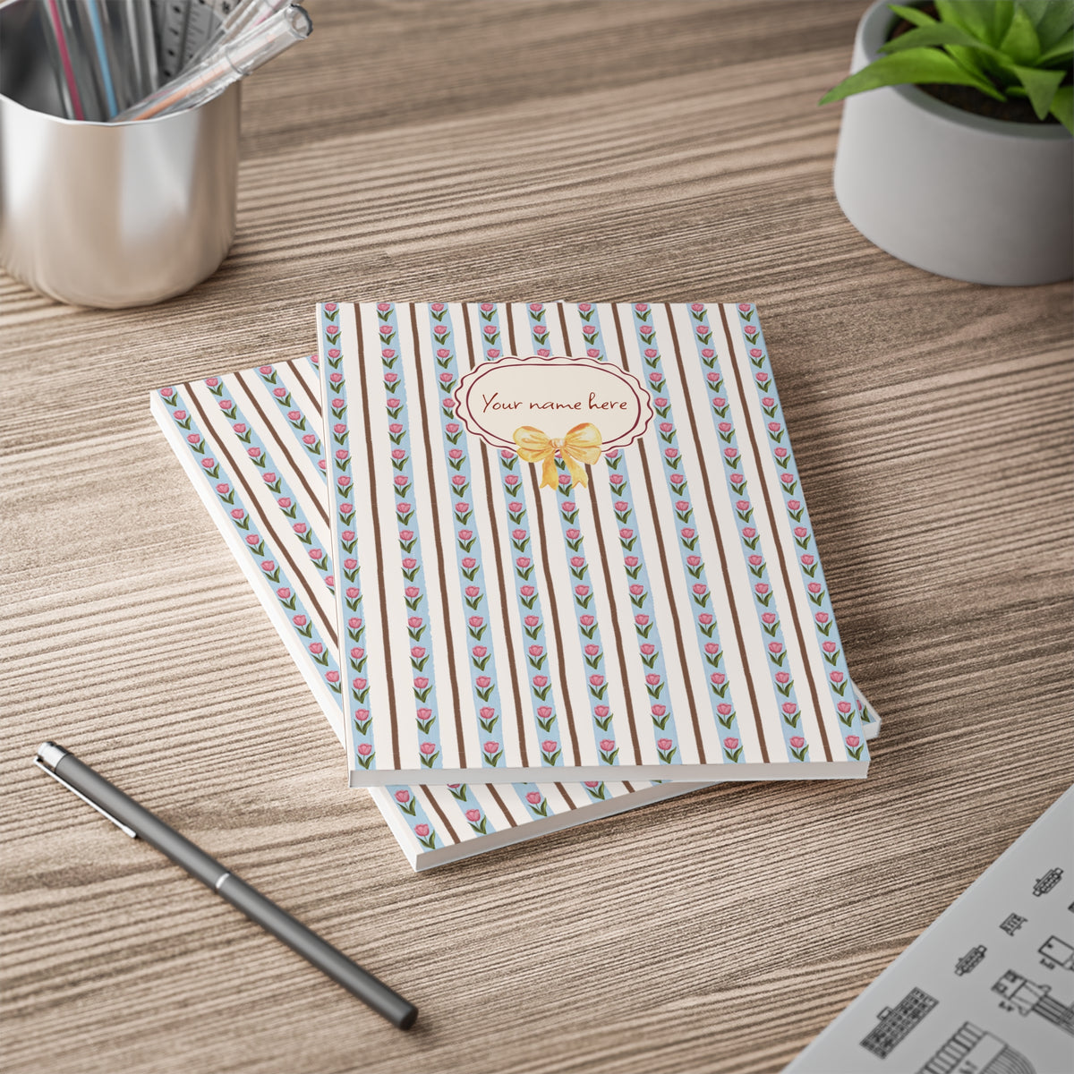 Two striped notebooks with a pink and green tulip pattern and a gold bow design on the cover, displayed on a wooden desk next to a pen, a cup of pens, and a small potted plant.