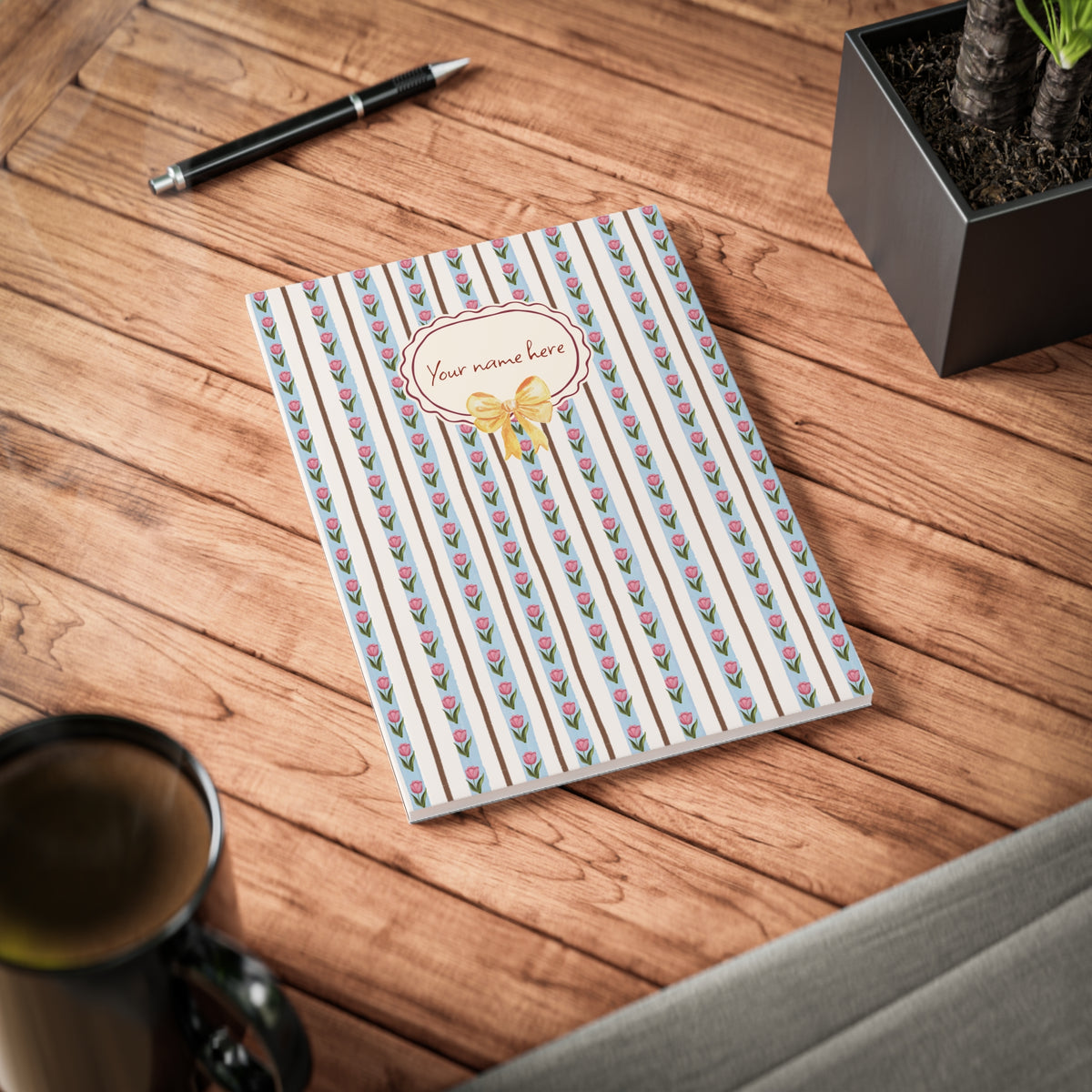 A personalized A5 softcover notebook with a vertical striped pattern of pink tulips on a white background, featuring a custom name label on the front, displayed on a wooden desk with a pen, mug, and plant.
