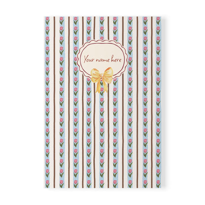 A Softcover A5 Notebook with a vertical striped pattern featuring pink tulips, brown stripes, and a central cream oval label with the text 'Your name here' and a yellow bow illustration, shown on a white background.