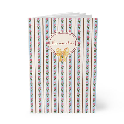 A personalized Softcover A5 Notebook with a vertical striped pattern featuring pink tulips, brown stripes, and a central cream-colored oval label with a gold bow and the text 'Your name here'.