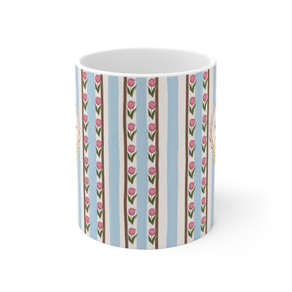 A white ceramic mug featuring a vertical striped pattern with light blue, beige, and brown stripes, decorated with pink tulip motifs and small yellow floral accents.