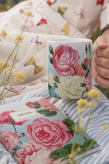 A white ceramic mug with a pink and cream floral print of a rose garden is held by a hand, set against a cozy background of a matching floral blanket and a blue-and-white striped pillow. A matching floral notebook lies in the foreground with a personalised label.